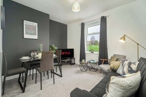 Una sala de estar moderna con una pared gris oscuro, un sofá negro, una mesa de comedor con sillas, un televisor mostrando Netflix y un amplio ventanal con cortinas.