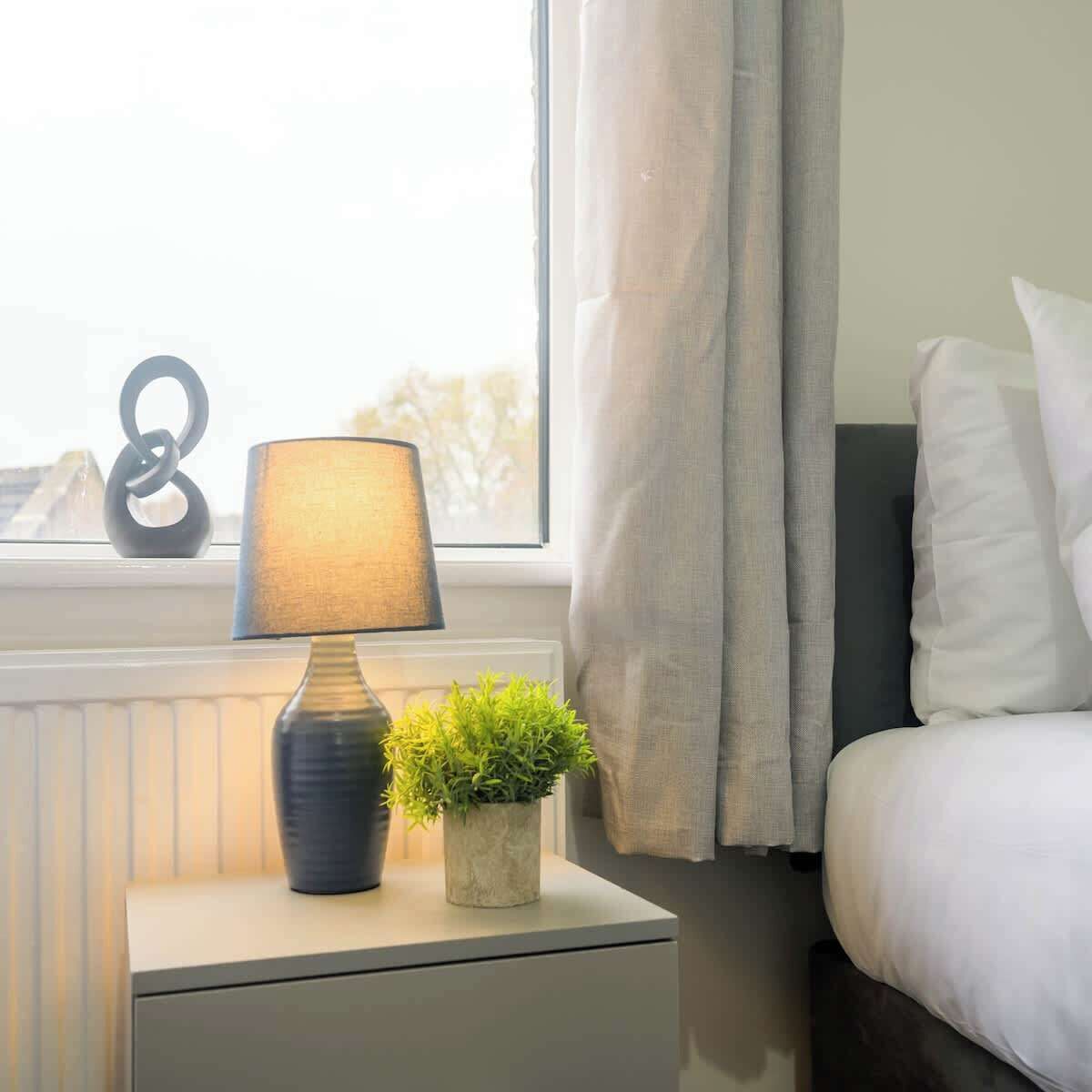 Un dormitorio ordenado con una mesita de noche que tiene una lámpara y una planta pequeña, una ventana con cortinas y una cama con almohadas.