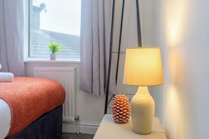 Una cama bien hecha con una colcha roja, una lámpara de noche blanca y una decoración de piña sobre una mesita de noche.