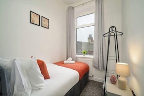 A hotel room with a bed, a window, a radiator, a lamp, and a coat rack.