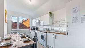 A modern kitchen with white cabinetry, a wooden dining table set for six, a washing machine, and a view of a brick building outside. - Lily House - 3 Bed WIFI & Parking Hull (Kingston upon Hull)