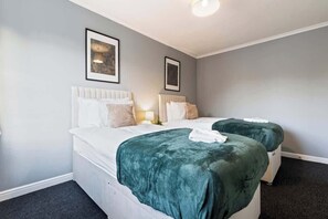 Une chambre d’hôtel avec deux lits, chacun doté d’une tête de lit blanche et d’une couverture verte. Des tableaux encadrés ornent le mur, et un luminaire est fixé au plafond.