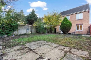 Un jardin à l’arrière d’une maison, avec une allée pavée, un grand arbre à feuilles persistantes et une maison en briques.