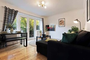 Ein modernes Wohnzimmer mit einem schwarzen Sofa, einem Esstisch mit Stühlen, einem an der Wand montierten Fernseher und einem großen Fenster mit Vorhängen.