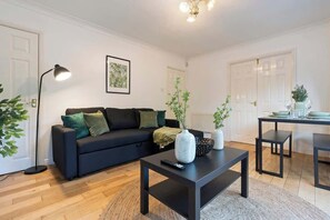 Ein modernes Wohnzimmer mit einem schwarzen Sofa, einem Couchtisch und einem Esstisch mit Stühlen. Es gibt eine Stehlampe, ein gerahmtes Bild an der Wand und einen Blumentopf.
