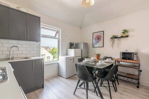 Et kompakt køkken med lysegrå skabe, et spisebord med stole, en mikrobølgeovn og et lille køleskab.