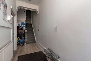 A narrow hallway with a staircase leading to a landing. There are bicycles and bags stored against the wall, and a black metal rack on the floor.
