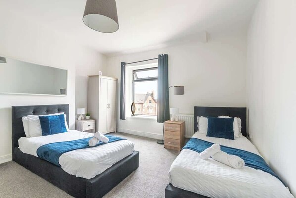 A bedroom with two beds, a window with curtains, a bedside table, and a lamp.