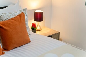 Een keurig opgemaakt bed met streepjesbeddengoed, een houten nachtkastje met een lamp en een kleine potplant.