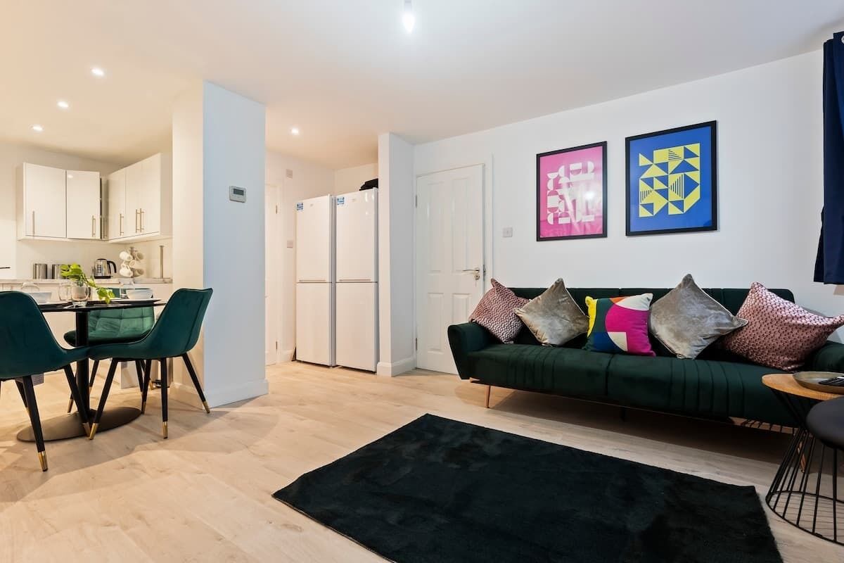 Un salon moderne avec un canapé vert, un coin repas avec des chaises vertes et une cuisine dotée de meubles blancs et d’un réfrigérateur.