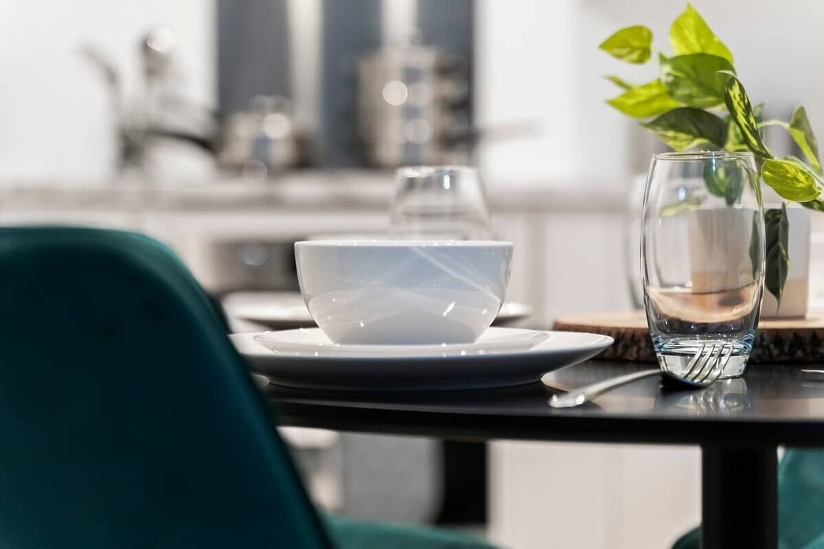 A dining table with a white bowl, a glass of water, and a fork. There is a green plant in a vase to the right.