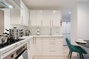 A modern kitchen with white cabinets, a stove, and a dining area with a green chair. - Ruby’s Retreat-4 Bed- Contractor Disc- WiFi (Hertfordshire)