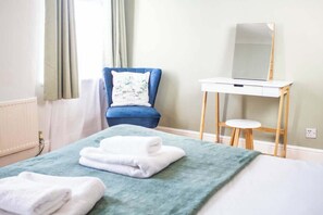 Ein Hotelzimmer mit Bett, blauem Sessel, kleinem Holztisch und Spiegel sowie einem Fenster mit Vorhängen.