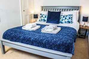 A neatly made bed with a blue textured bedspread, two folded white towels, and a variety of pillows including a dark blue one and patterned pillows.