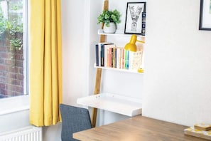 A room with a wooden table, a chair, a white shelf with books and a plant, and a window with yellow curtains.