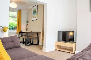 A living room with a sofa, a TV on a wooden stand, a dining table with chairs, and a window with curtains.