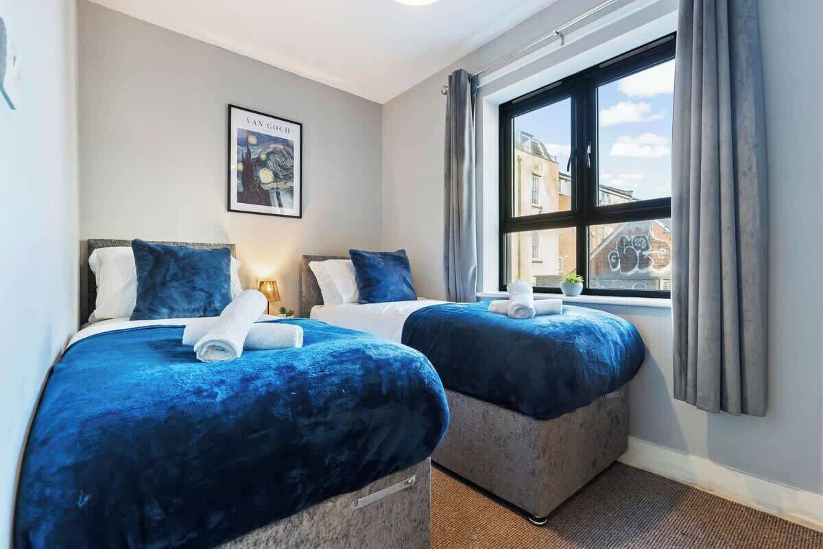 A hotel room with two beds, each with a blue bedspread and matching pillows. There is a framed picture on the wall, a window with curtains, and a view of buildings outside.