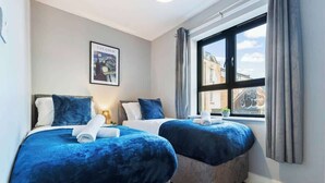 Ein Hotelzimmer mit zwei Betten, jeweils mit blauer Bettdecke und passenden Kissen. An der Wand hängt ein gerahmtes Bild, ein Fenster mit Vorhängen und Blick auf umliegende Gebäude.
