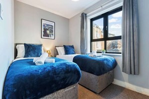 A hotel room with two beds, each with a blue bedspread and matching pillows. There is a framed picture on the wall, a window with curtains, and a view of buildings outside.