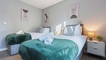 A hotel room with two beds, each with a green bedspread and white linens, a bedside table with a lamp, and artwork on the walls.