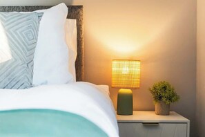 Una cama bien hecha con almohadas estampadas, una lámpara de noche y una pequeña planta en maceta sobre una mesita.