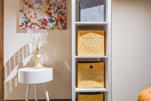 A room with a white lamp on a small table, a shelf with patterned cushions, and a floral painting on the wall. - Daffodil Apartment 4 Bedroom in Pontypool (Torfaen Principal Area)
