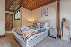 A bedroom with a wooden ceiling, a bed with a grey and orange blanket, a nightstand with a lamp, and a wooden door. - Daffodil Apartment 4 Bedroom in Pontypool (Torfaen Principal Area)