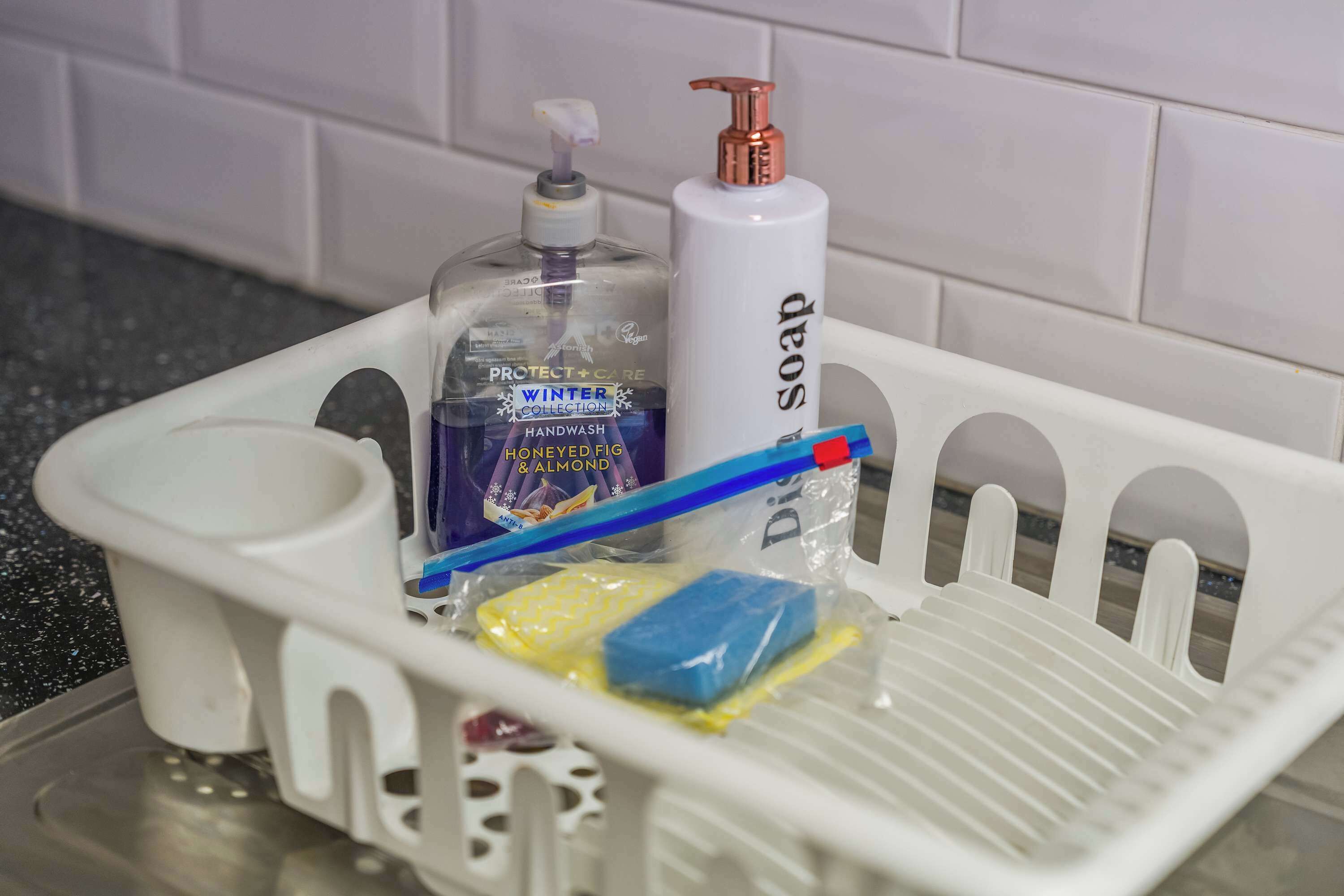 Un coin lavabo dans la salle de bain, avec un bac en plastique blanc, un distributeur de savon, un flacon de savon pour les mains, une brosse à dents bleue et une éponge jaune.