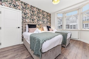 A bedroom with two beds, a patterned wallpaper, a white door, and a window with a view of buildings.