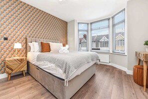 A modern bedroom with a geometric patterned wall, a large bed with a grey blanket, and a view of buildings through large windows.