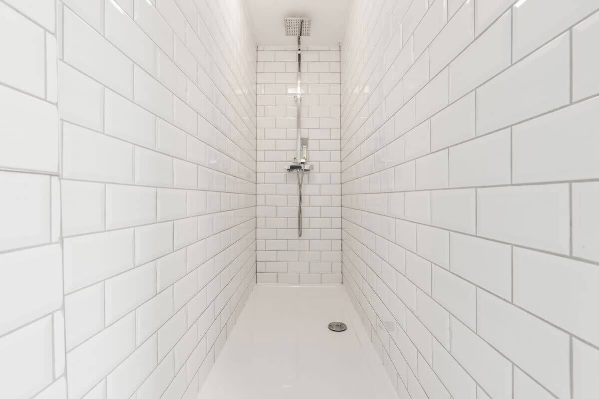 A narrow, white-tiled shower stall with a modern showerhead and a drain in the center.