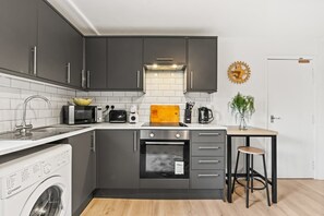 Une cuisine compacte avec des meubles de rangement sombres, un four à micro-ondes, un grille-pain, une machine à café et une planche à découper en bois.