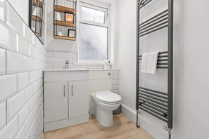 A compact bathroom with white tiled walls, a white toilet, a small window, and a towel rack.