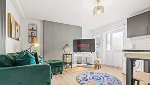 Una sala moderna con un sofá verde, un tapete redondo estampado, un televisor con el logotipo de Netflix, un taburete pequeño y una zona de cocina con armarios.