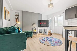 Een moderne woonkamer met een groene bank, een rond tapijt met een patroon, een televisie met Netflix-logo, een klein krukje en een keuken met kastjes.