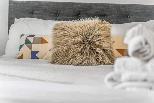 A bed with a tufted headboard, a geometric-patterned pillow, and a fluffy, shaggy pillow.