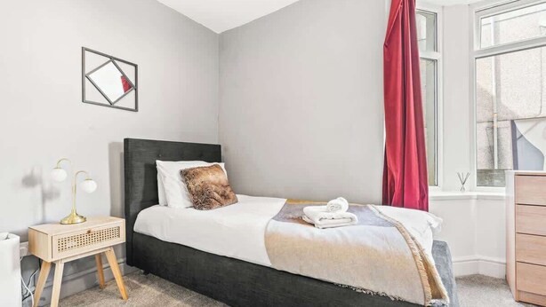 Un dormitorio con cama, mesita de noche, lámpara, ventana con cortinas y una obra de arte colgada en la pared.