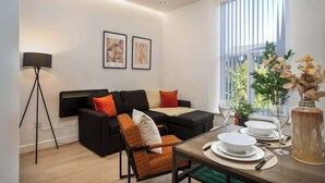 Een moderne woonkamer met een zwart modulaire bank, een eettafel voor twee personen en een groot raam met jaloezieën.