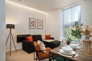 A modern living room with a black sectional sofa, a dining table set for two, and a large window with blinds.