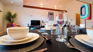 Una mesa de comedor dispuesta con platos, tazones, cubiertos y vasos, con una decoración moderna y un televisor al fondo.