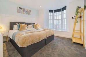 A modern bedroom with a large bed, a bedside table, a window with a view, and a wooden ladder with plants.