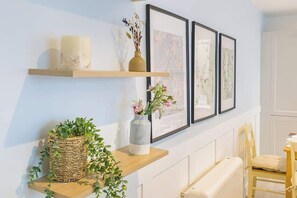 Une pièce avec une étagère en bois où sont posées des bougies et un vase rempli de fleurs, un panier en osier contenant des plantes vertes et des tableaux encadrés accrochés au mur.