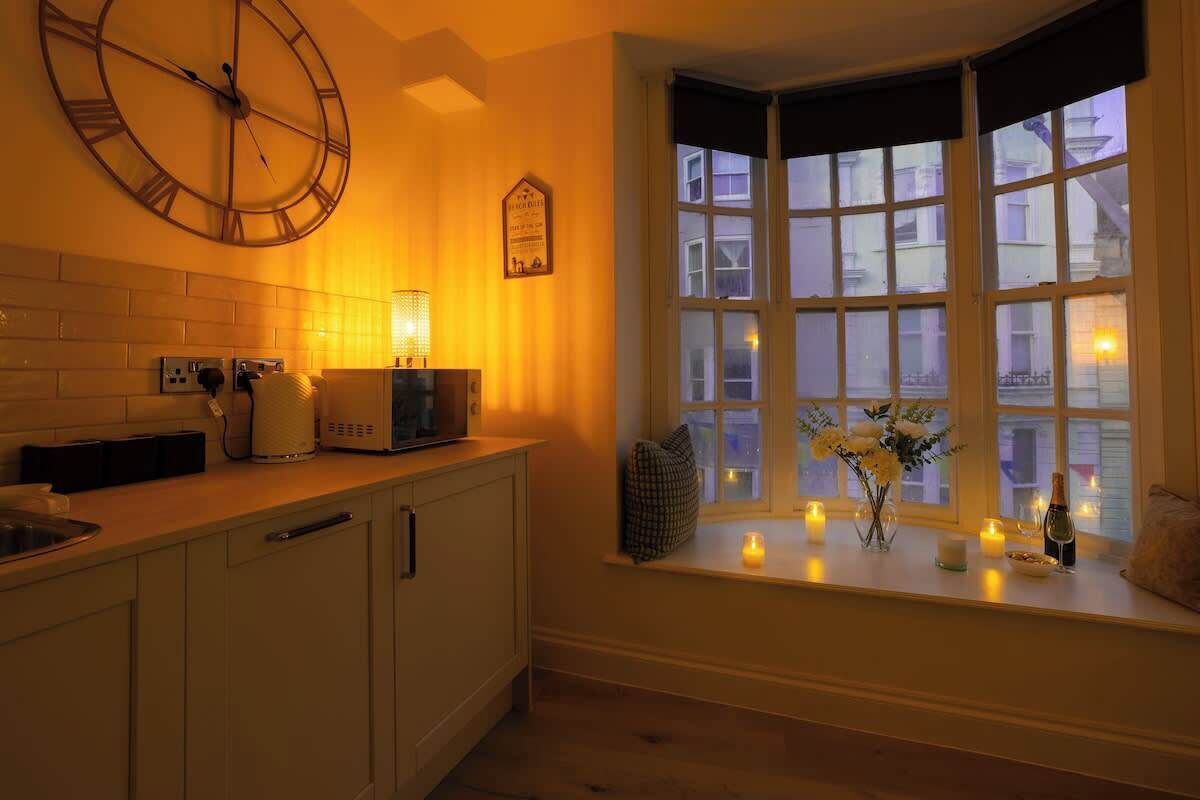 Una cocina con un reloj de pared grande, una ventana con vista a edificios y un estante con velas y un jarrón de flores.