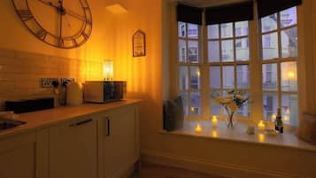 Una cocina con un reloj de pared grande, una ventana con vista a edificios y un estante con velas y un jarrón de flores.