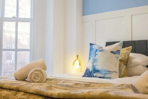 Un lit bien fait, avec une serviette roulée, une lampe de chevet et des oreillers, dans une pièce dotée d’une fenêtre et d’un mur bleu.