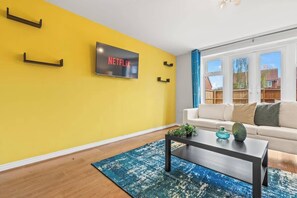 Un salon avec un mur jaune, un téléviseur à écran plat fixé au-dessus d’une table basse noire, et un canapé blanc agrémenté de coussins.