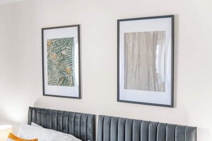 A bedroom with a grey upholstered headboard, a white pillow, and an orange accent pillow. Two framed artworks are hung on the wall above the bed.
