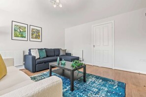Un salon moderne avec un canapé foncé, une table basse et un tapis à motifs bleus. Deux tableaux encadrés sont accrochés au mur, et une porte blanche est visible au fond.