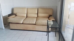 A beige leather sofa in a room with white tiled flooring, a glass table, and two framed pictures on the wall.