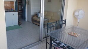 A room with a sliding glass door leading to a balcony, a small kitchenette, a sofa, and a table with a fan.